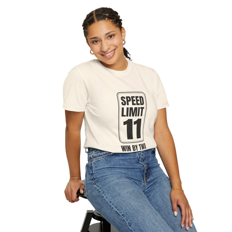 May include: A cream-colored short-sleeve t-shirt featuring a black graphic print of a rectangular road sign. The sign displays the text SPEED LIMIT 11 and WIN BY TWO in bold black lettering. The shirt is paired with blue denim jeans.
