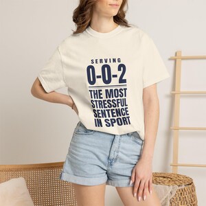 May include: A cream-colored short-sleeve t-shirt featuring dark blue text that reads: SERVING 0-0-2 THE MOST STRESSFUL SENTENCE IN SPORT. The shirt has a crew neckline and is paired with light blue denim shorts with a cuffed hemline.