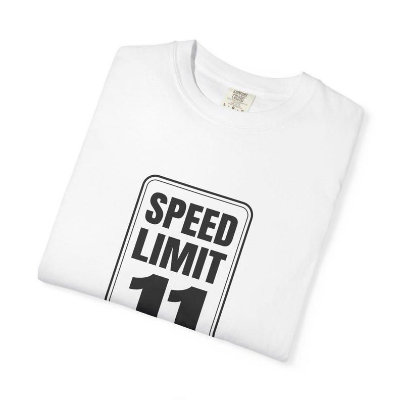 May include: A white short-sleeved t-shirt featuring a black graphic print of a rectangular road sign. The sign contains the text SPEED LIMIT in bold capital letters above the number 11. The shirt has a crew neckline and a visible interior neck label.