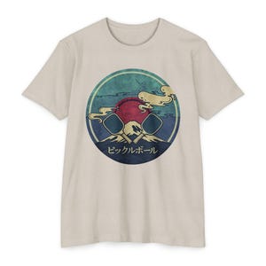 May include: A beige short-sleeve t-shirt featuring a circular graphic on the chest. The design displays two crossed pickleball paddles over a mountain silhouette, a large red sun, and stylised clouds. The Japanese text reads "Pickleball".