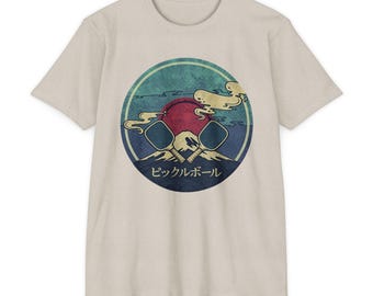 Op de afbeelding: Een beige t-shirt met korte mouwen en een ronde afbeelding op de borst. Het ontwerp toont twee gekruiste pickleball-peddels boven een bergsilhouet, een grote rode zon en gestileerde wolken. De Japanse tekst betekent "Pickleball".