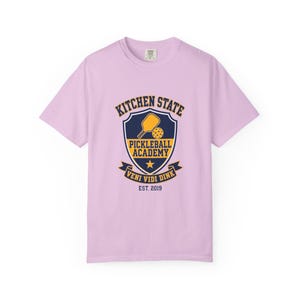 May include: A light purple short-sleeve t-shirt featuring a dark blue and yellow shield graphic on the chest. The design includes a pickleball paddle, a ball, a star, and the text KITCHEN STATE PICKLEBALL ACADEMY VENI VIDI DINK EST. 2019.