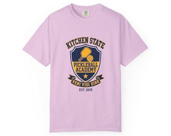 May include: A light purple short-sleeve t-shirt featuring a dark blue and yellow shield graphic on the chest. The design includes a pickleball paddle, a ball, a star, and the text KITCHEN STATE PICKLEBALL ACADEMY VENI VIDI DINK EST. 2019.