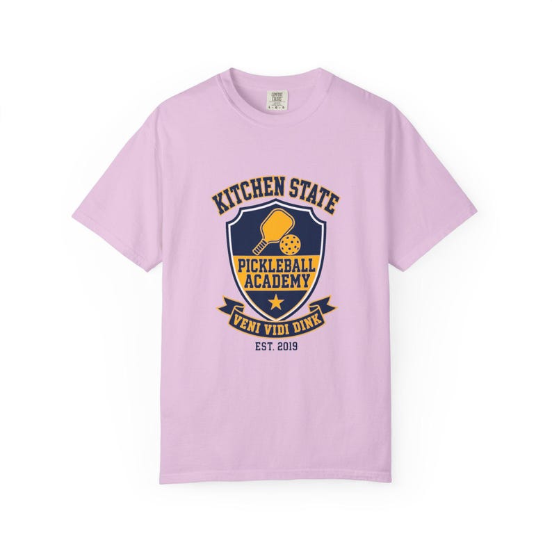 Może przedstawiać: Jasnofioletowy t-shirt z kr&oacute;tkim rękawem z granatowo-ż&oacute;łtym motywem tarczy na piersi. Wz&oacute;r zawiera rakietkę do pickleballa, piłkę, gwiazdę oraz napis KITCHEN STATE PICKLEBALL ACADEMY VENI VIDI DINK EST. 2019.