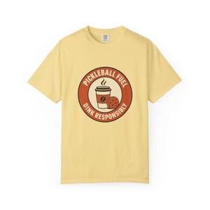 May include: A pale yellow short-sleeve t-shirt featuring a circular graphic on the chest. The graphic contains a coffee cup and a pickleball, surrounded by the text "PICKLEBALL FUEL" and "DINK RESPONSIBLY" in a bold, dark red circular border.