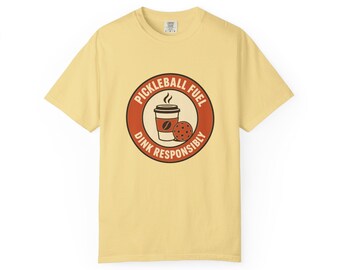 May include: A pale yellow short-sleeve t-shirt featuring a circular graphic on the chest. The graphic contains a coffee cup and a pickleball, surrounded by the text "PICKLEBALL FUEL" and "DINK RESPONSIBLY" in a bold, dark red circular border.