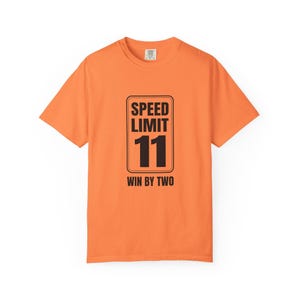May include: A bright orange short-sleeve t-shirt featuring a black graphic of a rectangular speed limit sign. The sign displays the text SPEED LIMIT 11 and the phrase WIN BY TWO in bold black lettering centered on the front of the garment.
