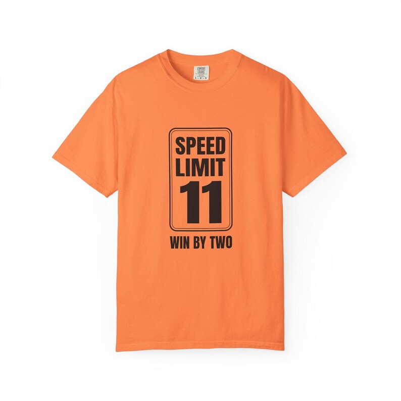 May include: A bright orange short-sleeve t-shirt featuring a black graphic of a rectangular speed limit sign. The sign displays the text SPEED LIMIT 11 and the phrase WIN BY TWO in bold black lettering centered on the front of the garment.