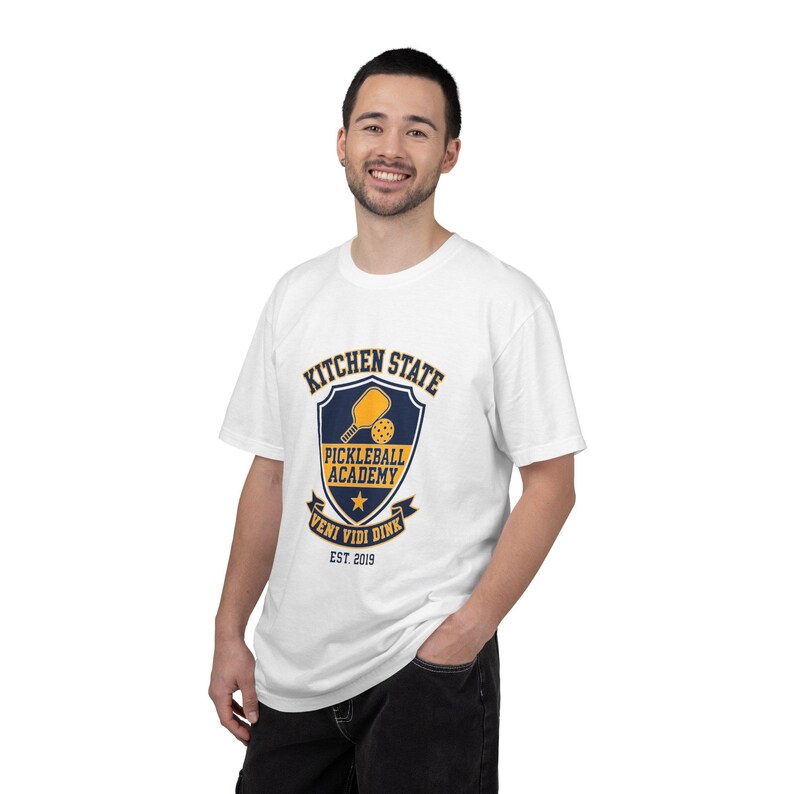Może przedstawiać: Biały t-shirt z kr&oacute;tkim rękawem z granatowo-ż&oacute;łtą grafiką tarczy na piersi. Wz&oacute;r zawiera rakietkę do pickleballa, piłkę, gwiazdę oraz napis Kitchen State Pickleball Academy Veni Vidi Dink Est. 2019.