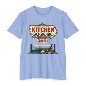 May include: A light blue t-shirt featuring a graphic of a vintage-style sign that reads KITCHEN CLOSED in red and dark blue lettering. Below the sign is a pickleball court illustration with a net, a yellow ball, and a black and red paddle.