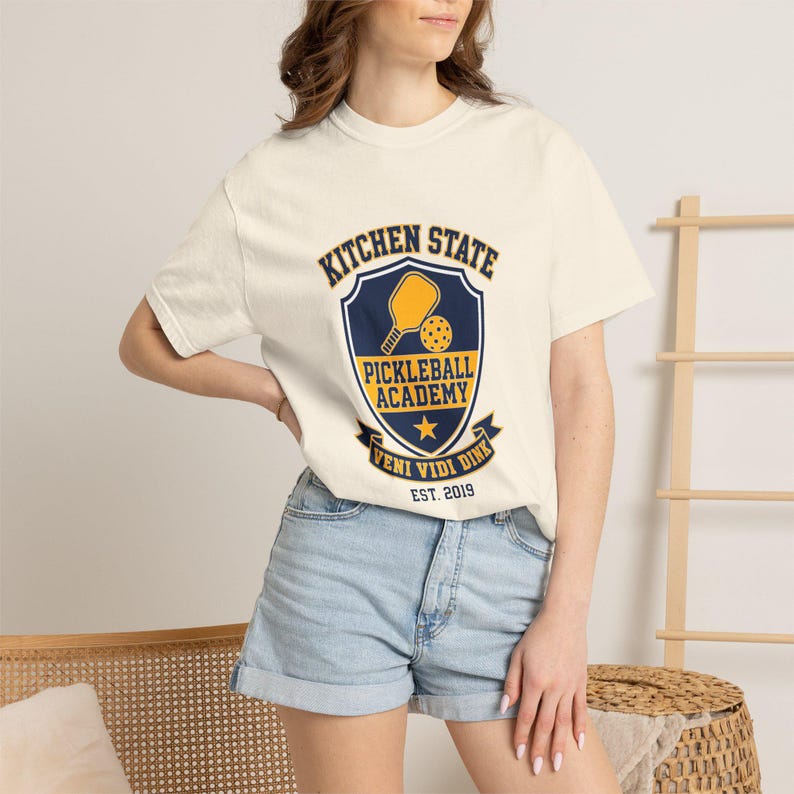 Może przedstawiać: Kremowy t-shirt z kr&oacute;tkim rękawem z granatowo-ż&oacute;łtą grafiką tarczy. Napis głosi KITCHEN STATE PICKLEBALL ACADEMY VENI VIDI DINK EST. 2019. Grafika zawiera ż&oacute;łtą rakietkę do pickleballa oraz ikonę ż&oacute;łtej piłeczki.