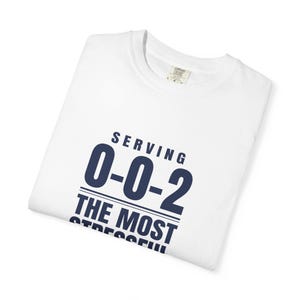 May include: A white short-sleeved t-shirt featuring dark blue text that reads "SERVING 0-0-2 THE MOST STRESSFUL" printed on the front. The shirt has a crew neckline and a visible brand label inside the collar area indicating a size large.