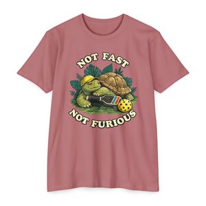 May include: A dusty rose t-shirt featuring a graphic of a green turtle wearing a yellow cap, holding a pickleball paddle next to a yellow pickleball, surrounded by green leaves, with the text NOT FAST NOT FURIOUS printed in cream.