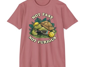 Op de afbeelding: Een oudroze t-shirt met een afbeelding van een groene schildpad met een gele pet, die een pickleball-peddel vasthoudt naast een gele pickleball, omringd door groene bladeren, met de tekst NOT FAST NOT FURIOUS in het crème gedrukt.