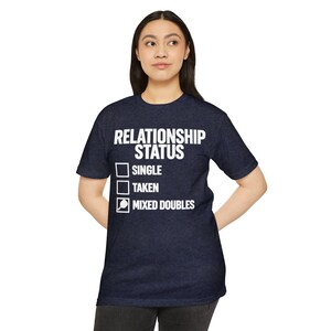Op de afbeelding: Een donkerblauw t-shirt met korte mouwen en witte tekst met de tekst RELATIONSHIP STATUS gevolgd door drie selectievakjes. De eerste twee zijn leeg, terwijl het derde vakje een wit pickleball-racketpictogram naast MIXED DOUBLES bevat.