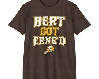 Op de afbeelding: Een donkerbruin t-shirt met korte mouwen en de tekst "BERT GOT ERNE'D" in een verweerd geel lettertype met een witte omlijning. Onder de tekst staat een kleine afbeelding van een geel-wit pickleball-racket en een gele pickleball-bal.