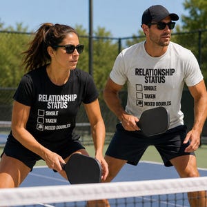 Op de afbeelding: Een zwart t-shirt en een lichtgrijs t-shirt, beide met de tekst RELATIONSHIP STATUS met selectievakjes voor SINGLE, TAKEN en een pickleball-paddle-icoon naast MIXED DOUBLES. Elk shirt wordt gedragen met een zwarte sportshort en een zonnebril.