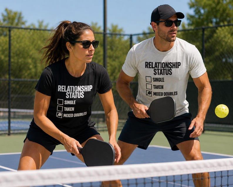 Op de afbeelding: Een zwart t-shirt en een lichtgrijs t-shirt, beide met de tekst RELATIONSHIP STATUS met selectievakjes voor SINGLE, TAKEN en een pickleball-paddle-icoon naast MIXED DOUBLES. Elk shirt wordt gedragen met een zwarte sportshort en een zonnebril.