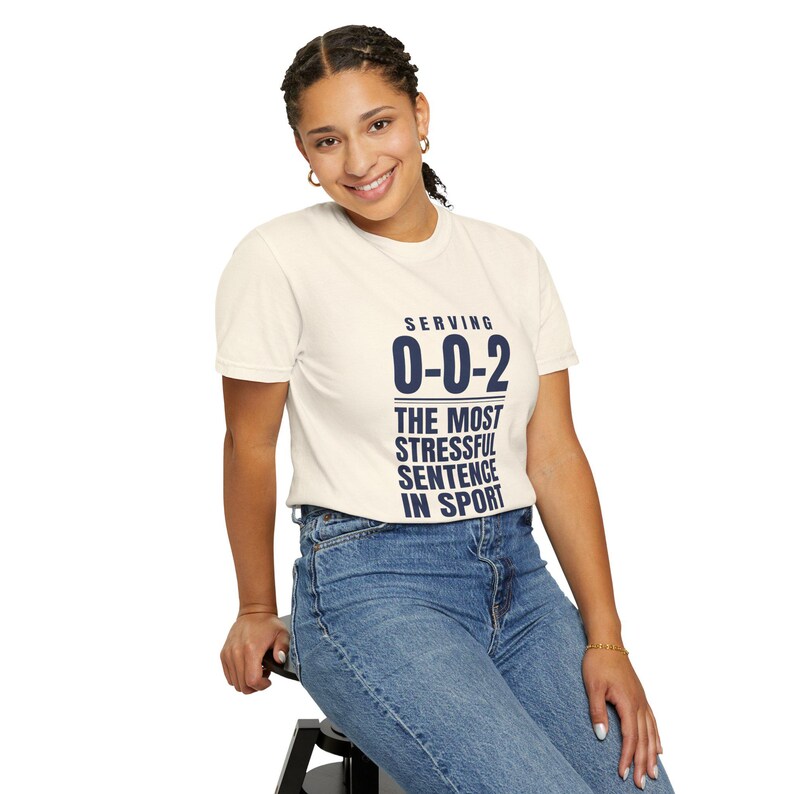 May include: A cream-colored short-sleeve t-shirt featuring dark blue text that reads: SERVING 0-0-2 THE MOST STRESSFUL SENTENCE IN SPORT. The shirt has a crew neckline and is paired with blue denim jeans, creating a casual and simple appearance.