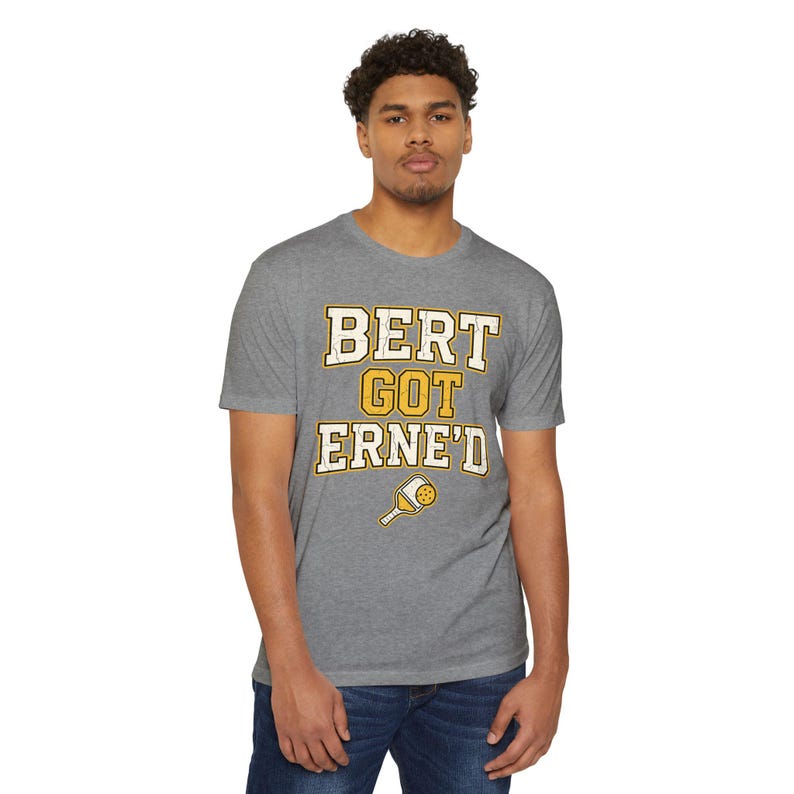 May include: A heather grey short-sleeve t-shirt featuring the text BERT GOT ERNE'D in a distressed yellow and white font. Below the text is a small graphic of a yellow pickleball paddle with a white handle and a dotted ball pattern.