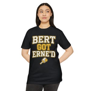 May include: A black short-sleeved t-shirt featuring the yellow and white text "BERT GOT ERNE'D" in a block font, accompanied by a small graphic of a yellow pickleball paddle with a white handle positioned below the text.