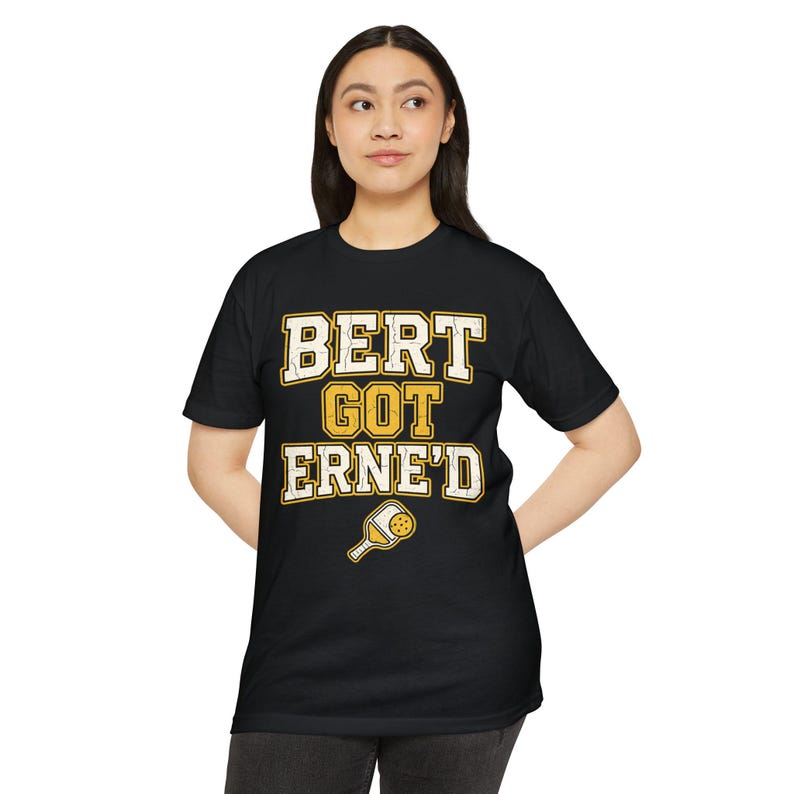 May include: A black short-sleeved t-shirt featuring the yellow and white text "BERT GOT ERNE'D" in a block font, accompanied by a small graphic of a yellow pickleball paddle with a white handle positioned below the text.