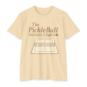 May include: A light beige short-sleeved t-shirt featuring a brown graphic print. The design includes the text The PickleBall Club above a line drawing of a pickleball court and a small ball. The shirt has a crew neckline and a standard fit.