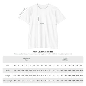 Op de afbeelding: Een wit t-shirt met korte mouwen en een maattabeldiagram dat breedte, lengte en mouwlengte aangeeft. Hieronder staat een tabel met Next Level 6210-maten in inches en centimeters, met een vermelding van productietoleranties van 3,8 cm.