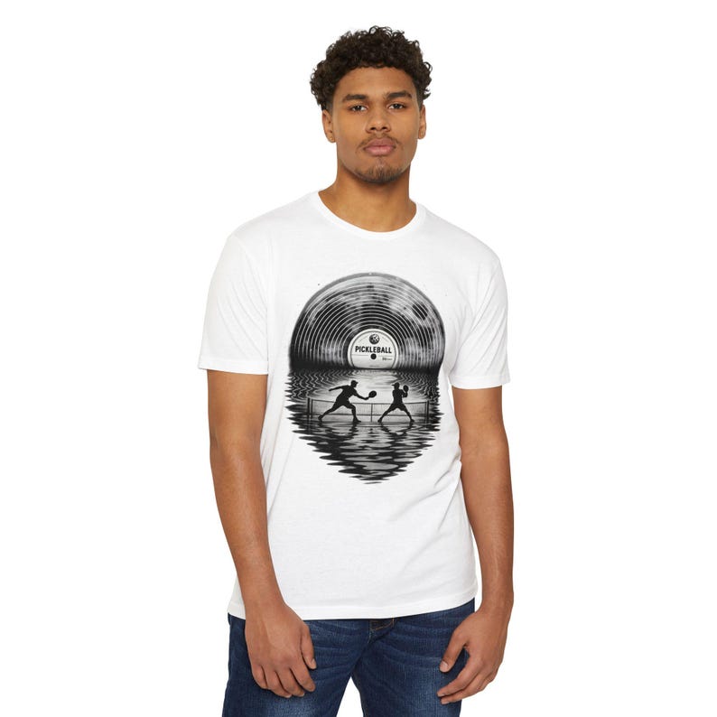 May include: A white short-sleeve t-shirt featuring a black and white graphic of a vinyl record that transitions into a pickleball court scene. The centre label of the record displays the text PICKLEBALL, with silhouettes of two players in action.