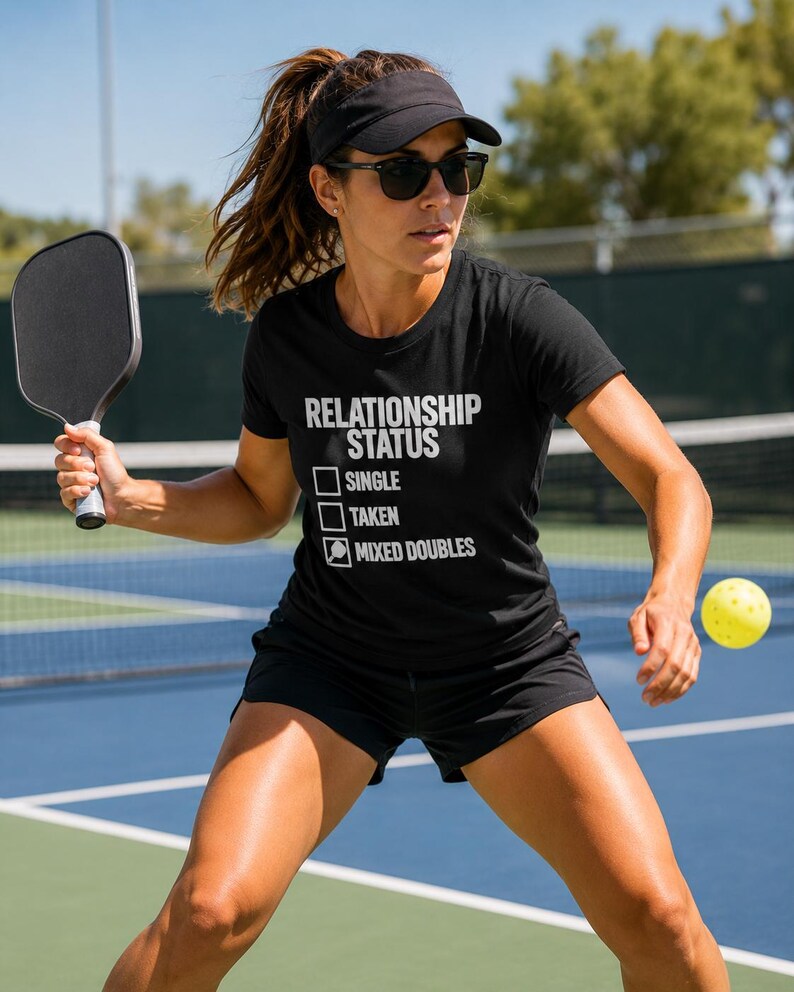 Op de afbeelding: Een zwart T-shirt met korte mouwen en witte tekst RELATIONSHIP STATUS met drie selectievakjes. De eerste twee vakjes zijn leeg, terwijl het derde vakje een icoon van een pickleball-racket bevat naast de tekst MIXED DOUBLES.