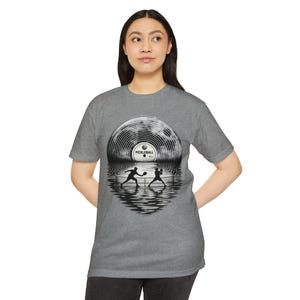 May include: A heather grey short-sleeve t-shirt featuring a graphic print of two pickleball players in silhouette on a court, with their reflections below and a large vinyl record shaped like a full moon in the background labelled PICKLEBALL.
