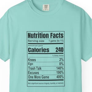 May include: A light mint green t-shirt featuring a black graphic print of a Nutrition Facts label. The text lists serving size as 1 game, calories as 240, and percentages for Knees, Ego, Trash Talk, Excuses, and One More Game, with a bottom disclaimer.