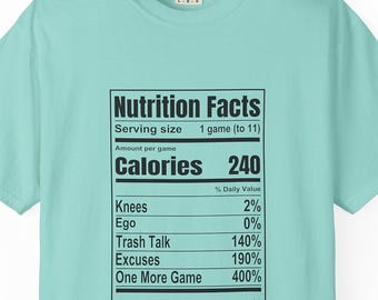 May include: A light mint green t-shirt featuring a black graphic print of a Nutrition Facts label. The text lists serving size as 1 game, calories as 240, and percentages for Knees, Ego, Trash Talk, Excuses, and One More Game, with a bottom disclaimer.