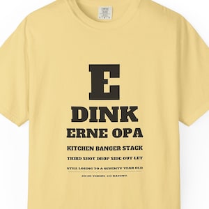 May include: A pale yellow short-sleeve t-shirt featuring a black graphic resembling an eye chart. The text reads E, DINK, ERNE OPA, KITCHEN BANGER STACK, THIRD SHOT DROP SIDE OUT LET, STILL LOSING TO A SEVENTY YEAR OLD, and 20/20 VISION. 3.0 RATING.