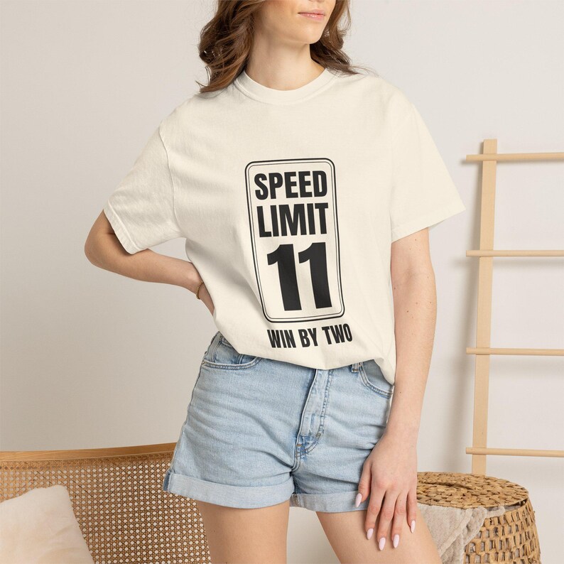 May include: A cream-colored short-sleeve t-shirt featuring a black graphic of a rectangular speed limit sign. The sign displays the text SPEED LIMIT 11 and WIN BY TWO in bold black lettering. The shirt is paired with light blue denim shorts.