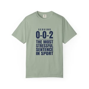 May include: A sage green short-sleeve t-shirt featuring dark navy blue text that reads: SERVING 0-0-2 THE MOST STRESSFUL SENTENCE IN SPORT. The shirt has a crew neckline and a standard fit, with the text centered on the front chest area.
