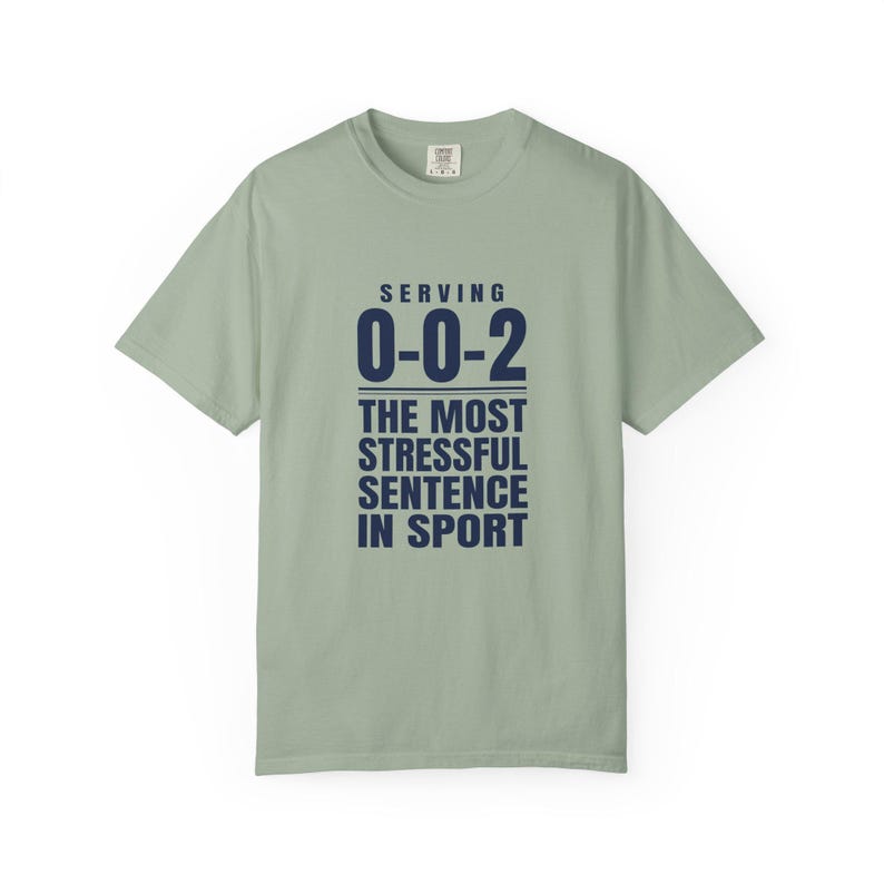 May include: A sage green short-sleeve t-shirt featuring dark navy blue text that reads: SERVING 0-0-2 THE MOST STRESSFUL SENTENCE IN SPORT. The shirt has a crew neckline and a standard fit, with the text centered on the front chest area.