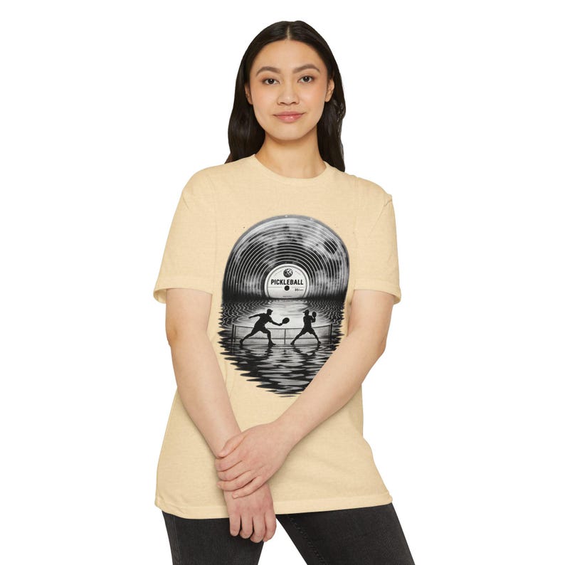 May include: A light tan short-sleeve t-shirt featuring a graphic print of a vinyl record shaped like a full moon above a water surface. The design shows two silhouettes playing pickleball on a court, with the text PICKLEBALL on the record label.