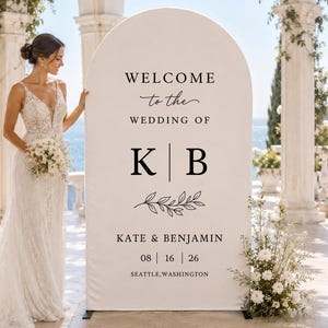 May include: An arched wedding welcome sign featuring black text on a white background. The text reads: WELCOME to the WEDDING OF K | B, KATE & BENJAMIN, 08 | 16 | 26, SEATTLE, WASHINGTON. A black line drawing of a leafy branch is centred below the initials.