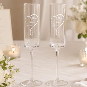 May include: Two clear glass champagne flutes featuring an etched design of a heart with a swirling tail. The text "Israel & Stevie" and the date "08.11.2024" are engraved inside the heart shape on each flute, which has a tall, slender bowl.