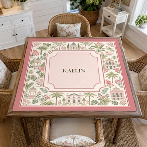 May include: A square table cover featuring a central cream-coloured panel with the name KAELIN printed in dark text. The border displays a repeating pattern of pink buildings, green palm trees, and tropical foliage on a soft pink background.