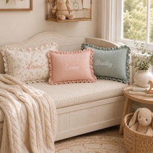 May include: Three rectangular throw pillows with ruffled edges, each featuring embroidered names. One pillow is cream with a floral pattern, one is solid pink with the name Emma, and one is solid sage green with the name Bradley.