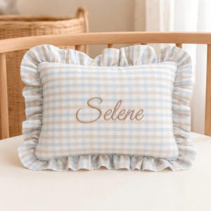 May include: A rectangular throw cushion featuring a light blue and beige gingham check pattern with a ruffled border. The name Selene is embroidered in a brown cursive script across the centre of the cushion, which is made of a soft fabric.