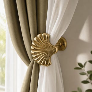 May include: A gold-toned metal curtain tieback shaped like a scalloped seashell. The decorative hardware features a polished, reflective finish and is mounted to a wall, holding back a thick olive green curtain and a sheer white curtain panel.