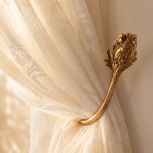 Può includere: Un fermatenda in metallo color oro con un terminale decorativo ornato a forma di foglia. Il gancio curvo sostiene un pannello di tenda trasparente color bianco sporco con un delicato motivo floreale in pizzo che lascia filtrare la luce.