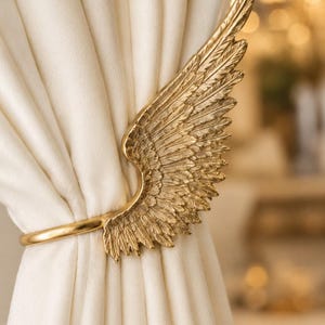 May include: A gold-colored metal curtain tieback featuring a detailed, textured wing design with individual feathers. The curved metal arm secures a gathered, off-white fabric curtain, holding the material in place with a smooth, polished finish.