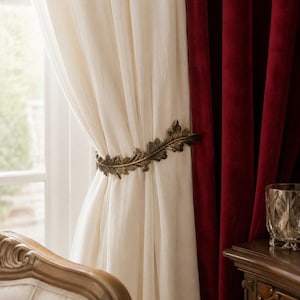 May include: A metallic bronze-toned curtain tieback shaped like a long, detailed oak leaf branch. It is wrapped around a cream-colored sheer curtain panel, positioned next to a heavy, deep red velvet curtain hanging in front of a window.