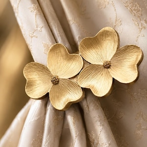 Puede incluir: Un alzapaños de cortina de metal en tono dorado que presenta dos formas de flores de tres pétalos con una textura cepillada y un pequeño centro circular texturizado. El accesorio floral está sujeto alrededor de una cortina de tela color crema claro con un patrón floral sutil.