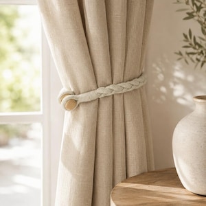 May include: A beige linen-textured curtain held back by a braided off-white rope tieback. The tieback features two smooth, light-coloured wooden circular toggles at the fastening point, securing the fabric in vertical folds.