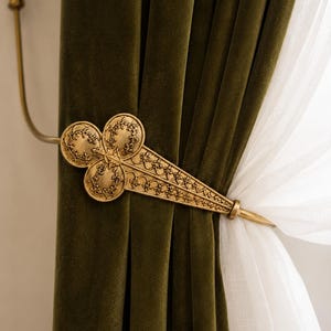 May include: A gold-toned metal curtain tieback featuring an ornate, clover-shaped design with intricate filigree patterns. The tieback holds back a heavy, dark olive green velvet curtain alongside a sheer, lightweight white curtain panel.